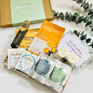 May include: A gift box filled with self-care items, including face masks, bath products, candles, and a handwritten note that says "Just a little something, we hope it makes you smile!"