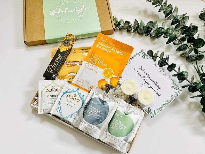 May include: A gift box filled with self-care items, including face masks, bath products, candles, and a handwritten note that says "Just a little something, we hope it makes you smile!"