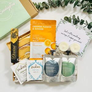 May include: A gift box filled with self-care items, including face masks, bath products, and candles. The box is decorated with white tissue paper and a handwritten note that says "Just a little something, we hope it makes you smile!"