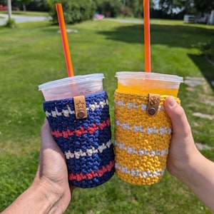 May include: Two colorful crocheted beverage holders with a striped pattern. The holder on the left is blue, white, and red. The holder on the right is yellow, white, and a light brown. Both holders have a brown leather strap with a star.