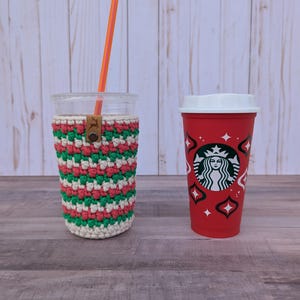 May include: A clear glass with an orange straw and a crocheted cozy in red, green, and white. Next to it is a red Starbucks cup with a white lid and logo.