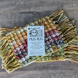 Puede incluir: Alfombra de taza hecha a mano con un diseño colorido en tonos amarillos, naranjas, rojos y verdes. La alfombra tiene un borde de flecos y una etiqueta que dice "alfombra de taza hecha a mano" con información adicional sobre el tamaño y las instrucciones de cuidado.