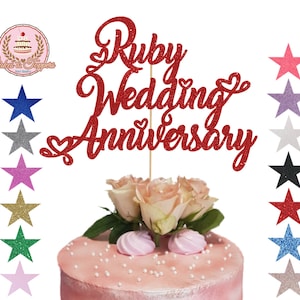 May include: Red glitter cake topper with the text "Ruby Wedding Anniversary". The topper is on a pink cake with white frosting and pearl sprinkles. The cake is decorated with pink roses. The image also shows a variety of star-shaped cake toppers in different colours.