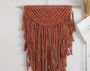 May include: A terracotta-colored macrame wall hanging with a half-circle design and long fringe. The hanging is made with thick cotton cord and is attached to a wooden dowel.