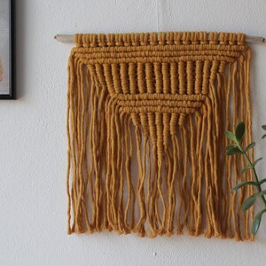 May include: A mustard yellow macrame wall hanging with a triangular pattern and fringe. The hanging is attached to a wooden dowel.