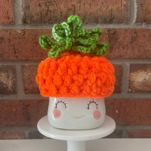 Op de afbeelding: Een witte keramische mok met een glimlachend gezicht en roze wangen, die een gehaakte oranje pompoenmuts met een groene steel draagt.