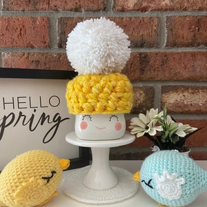 May include: A white ceramic mug with a yellow and white crocheted hat and a white pom pom on top. The mug has a happy face painted on it. There are two crocheted birds, one yellow and one blue, on either side of the mug.