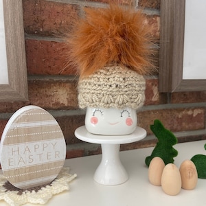 May include: A white ceramic mug with a happy face, wearing a crocheted hat with a brown pom-pom. The mug is on a white cake stand. There is a wooden sign that says "Happy Easter" and wooden eggs on a white surface.