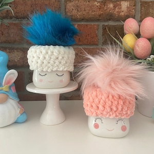 May include: Two white ceramic mugs with smiling faces and closed eyes. Each mug has a crocheted hat with a fluffy pom-pom on top. The hat on the left mug is white with a blue pom-pom, and the hat on the right mug is pink with a pink pom-pom.