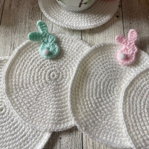 May include: White crocheted coasters with a scalloped edge and a bunny design. The coasters are decorated with small, colorful bunnies.