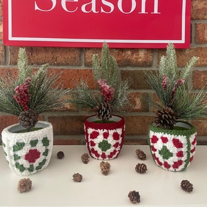 May include: Three crocheted plant pots with a red, white, and green pattern. The pots are filled with small pine trees and red berries. The pots are sitting on a white surface in front of a brick wall. A red sign with the text 'Tis the Season' is hanging on the wall.