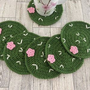 以下が含まれることがあります： 白いステッチとピンクの花のアクセントが付いた緑色のクロシェのコースター6枚。コースターは明るい木製の表面に円形に配置されています。