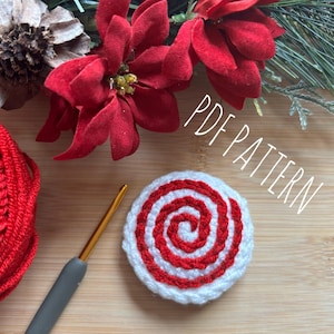 May include: A crochet candy cane ornament with red and white stripes. The ornament is on a wooden surface with a ball of red yarn, a crochet hook, and a sprig of red poinsettia flowers. The text "PDF PATTERN" is written in white on a black background.