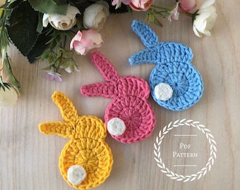 May include: Three crocheted bunny shapes in blue, pink, and yellow. Each bunny has a white circle for a tail. The bunnies are on a wooden surface with a floral arrangement in the background. The text "PDF PATTERN" is in a wreath.
