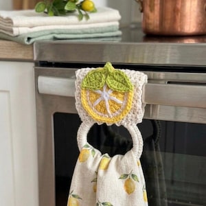 May include: A crochet towel holder featuring a yellow and white lemon slice motif with green leaves, attached to a beige ring. The holder secures a cream-colored kitchen towel printed with a repeating pattern of yellow lemons and green leaves.