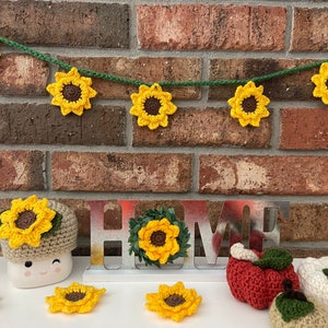May include: A crocheted sunflower garland with a brown center and yellow petals hangs on a brick wall.  The garland is strung with green yarn.  Below the garland is a metal sign that reads "HOME" with a crocheted sunflower wreath on the "O".  There are also crocheted sunflowers, apples, and pumpkins on a white surface.