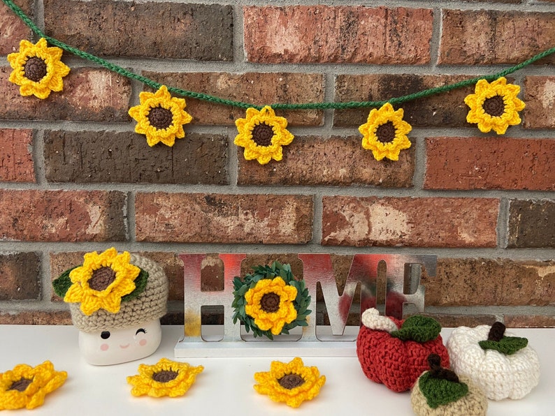 May include: A crocheted sunflower garland with a brown center and yellow petals hangs on a brick wall.  The garland is strung with green yarn.  Below the garland is a metal sign that reads "HOME" with a crocheted sunflower wreath on the "O".  There are also crocheted sunflowers, apples, and pumpkins on a white surface.