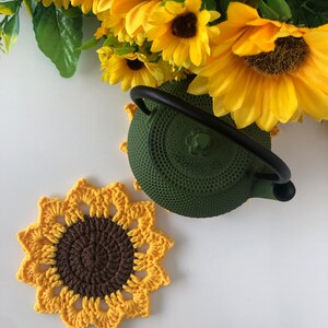 May include: A crocheted sunflower coaster in yellow and brown with a green cast iron teapot on top.