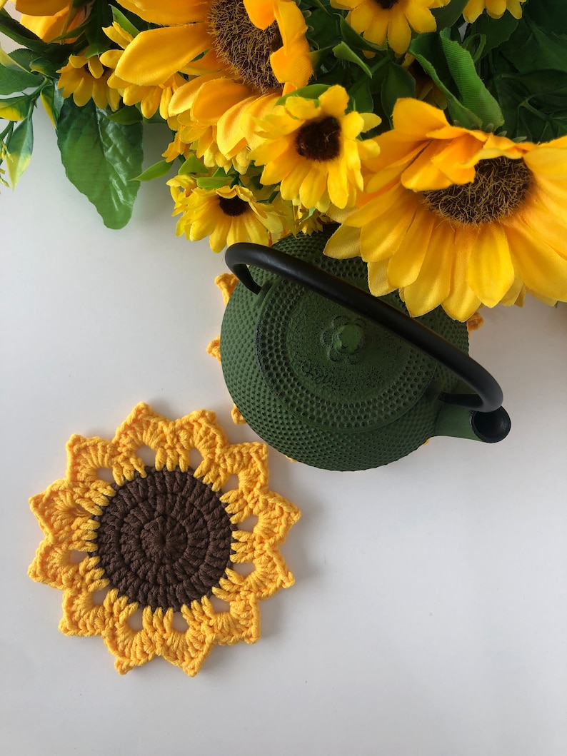 May include: A crocheted sunflower coaster in yellow and brown with a green cast iron teapot on top.