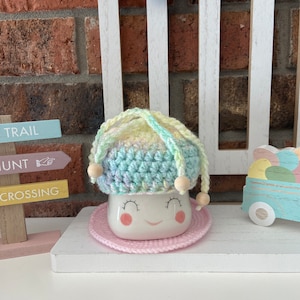 May include: A white ceramic mug with a smiling face, wearing a crocheted hat with rainbow colours and wooden beads. The mug is sitting on a pink crocheted coaster.