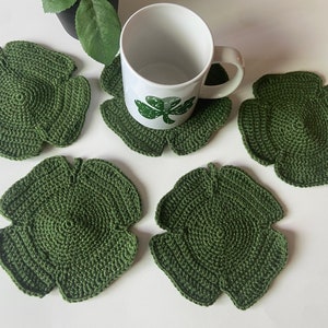 Puede incluir: Cuatro posavasos de ganchillo verde en forma de trébol de cuatro hojas. Los posavasos están sobre una superficie blanca. Una taza blanca con un diseño de trébol verde está sentada sobre uno de los posavasos.