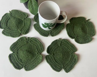May include: Four green crocheted coasters shaped like four-leaf clovers. The coasters are on a white surface. A white mug with a green shamrock design is sitting on one of the coasters.
