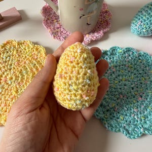 May include: A yellow crocheted egg is held in a hand, with a yellow and a teal crocheted coaster nearby. A mug with a floral design sits on a pink crocheted coaster. The items are on a white surface.