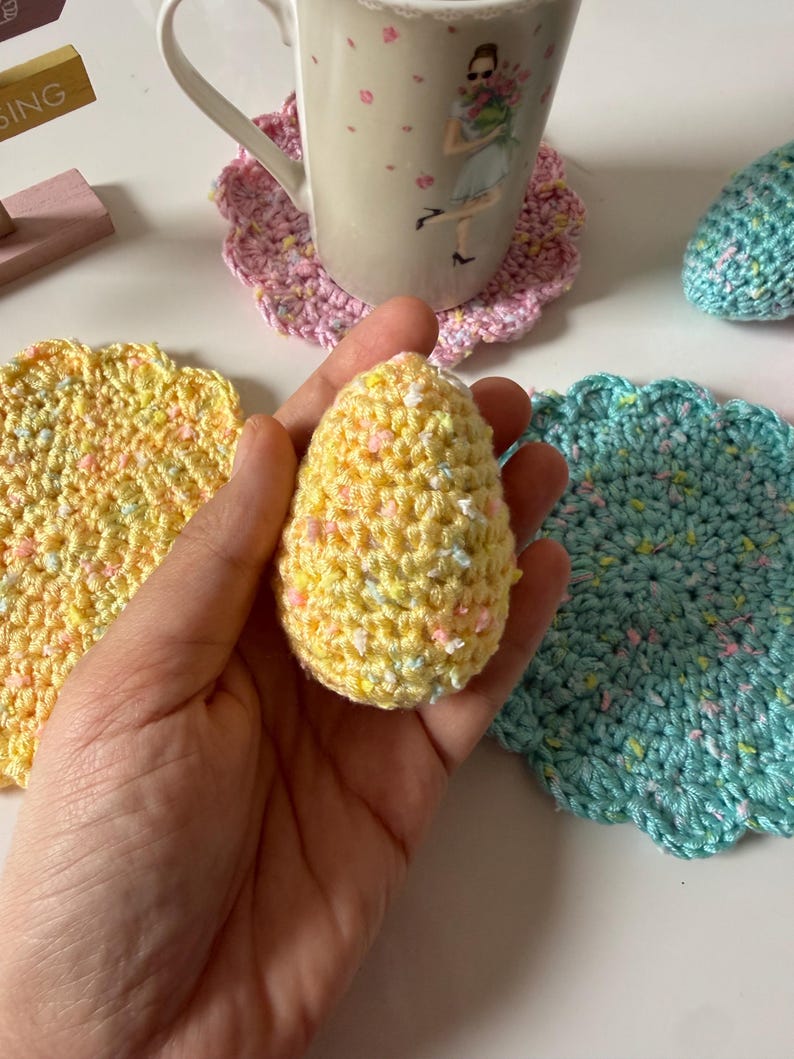 May include: A yellow crocheted egg is held in a hand, with a yellow and a teal crocheted coaster nearby. A mug with a floral design sits on a pink crocheted coaster. The items are on a white surface.