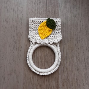 Puede incluir: Un soporte para toallas de ganchillo color crema que presenta una secci&oacute;n superior rectangular con borde festoneado y una base de anillo circular. Una aplicaci&oacute;n texturizada en forma de lim&oacute;n amarillo con una hoja verde oscuro est&aacute; unida al centro.