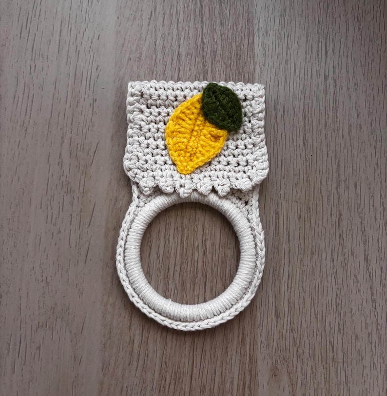 Puede incluir: Un soporte para toallas de ganchillo color crema que presenta una secci&oacute;n superior rectangular con borde festoneado y una base de anillo circular. Una aplicaci&oacute;n texturizada en forma de lim&oacute;n amarillo con una hoja verde oscuro est&aacute; unida al centro.
