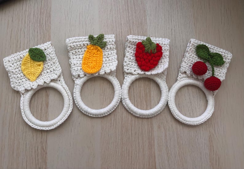 Puede incluir: Cuatro soportes para toallas de ganchillo con anillos blancos y partes superiores cuadradas con apliques de frutas. Los dise&ntilde;os incluyen un lim&oacute;n amarillo con hoja verde, una pi&ntilde;a naranja, una fresa roja y dos cerezas rojas con hojas verdes.