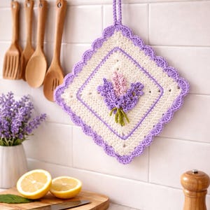 Puede incluir: Un agarrador de cocina cuadrado de ganchillo con un centro color crema y una aplicación de flores de lavanda tridimensional en tonos morados y verdes. El agarrador tiene un borde festoneado morado y un lazo a juego para colgarlo.