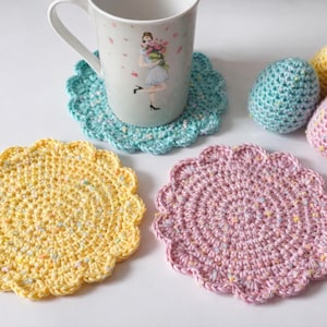 May include: A collection of handmade crochet items, including three coasters in yellow, pink, and teal, and three decorative egg-shaped items. A white mug with a floral design sits on a teal coaster. The items are arranged on a white surface.