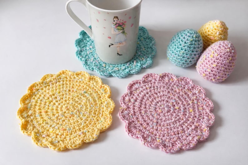 May include: A collection of handmade crochet items, including three coasters in yellow, pink, and teal, and three decorative egg-shaped items. A white mug with a floral design sits on a teal coaster. The items are arranged on a white surface.