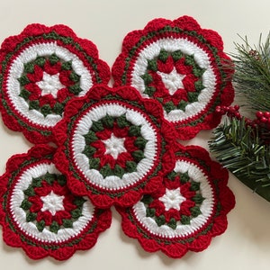 Peut inclure: Cinq dessous-de-plat en crochet rouge, blanc et vert avec un motif d'&eacute;toile au centre. Les dessous-de-plat sont dispos&eacute;s en cercle sur une surface blanche.