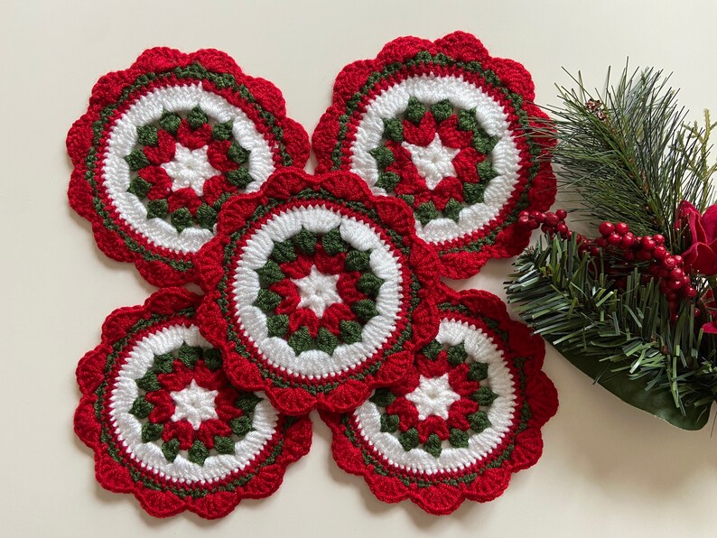 Peut inclure: Cinq dessous-de-plat en crochet rouge, blanc et vert avec un motif d'&eacute;toile au centre. Les dessous-de-plat sont dispos&eacute;s en cercle sur une surface blanche.