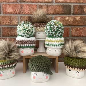 Puede incluir: Seis tazas de cerámica blanca con diferentes sombreros de ganchillo verde, marrón y blanco. Cada taza tiene una cara sonriente con los ojos cerrados.