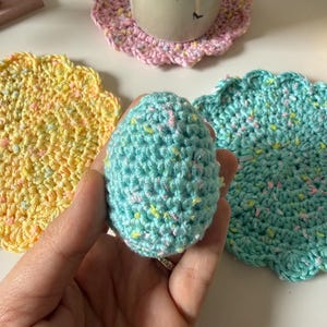 May include: A hand-crocheted, speckled, light blue egg is held near three crocheted coasters. The coasters are in shades of pink, yellow, and teal. A mug with a floral design sits on the pink coaster. The coasters have a scalloped edge.