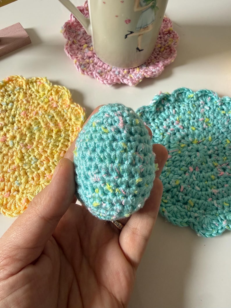 May include: A hand-crocheted, speckled, light blue egg is held near three crocheted coasters. The coasters are in shades of pink, yellow, and teal. A mug with a floral design sits on the pink coaster. The coasters have a scalloped edge.