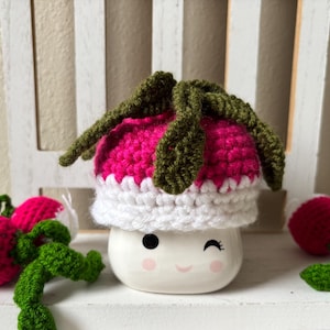 Peut inclure: Une tasse en céramique fantaisiste avec un visage qui cligne de l'œil, surmontée d'un chapeau de radis crocheté. Le chapeau est rose avec des feuilles vertes et un bord blanc. Des radis crochetés avec des tiges vertes sont éparpillés autour de la tasse.