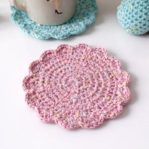 May include: A handmade, pink crochet coaster with a scalloped edge. The coaster is circular with a spiral pattern and flecks of yellow and blue. A light blue crochet coaster and a mug are visible in the background.