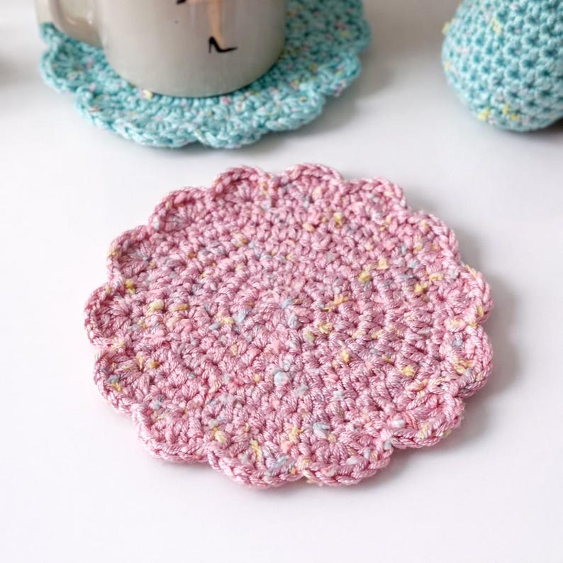 May include: A handmade, pink crochet coaster with a scalloped edge. The coaster is circular with a spiral pattern and flecks of yellow and blue. A light blue crochet coaster and a mug are visible in the background.