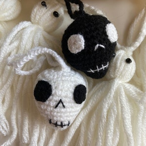 May include: A set of crocheted Halloween decorations featuring three white ghosts and one black skull. The ghosts have long white yarn tails and black eyes. The skull has white eyes and a stitched mouth.