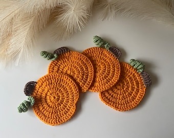 Puede incluir: Cuatro posavasos de ganchillo en forma de calabaza de color naranja con tallos verdes. Los posavasos están dispuestos en círculo sobre una superficie blanca.