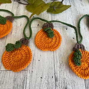 May include: A crocheted garland with five orange pumpkins with green stems and brown tops. The garland is strung together with green yarn.