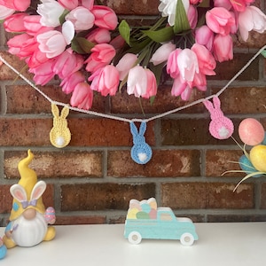 Può includere: Una ghirlanda di decorazioni a forma di coniglietto all'uncinetto in vari colori pastello, infilate su un cordoncino bianco. La ghirlanda è ambientata sullo sfondo di una ghirlanda di tulipani rosa e bianchi e un muro di mattoni. Decorazioni pasquali.