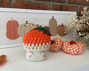May include: A white ceramic mug with a smiling face and a crocheted orange pumpkin hat. The mug is sitting on a white surface with two orange and white gingham pumpkins.