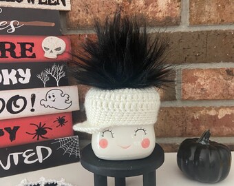 May include: White ceramic mug with a smiling face, wearing a white crocheted hat with a black fur pom-pom. The mug is sitting on a black stool with a white crocheted spiderweb on the floor.