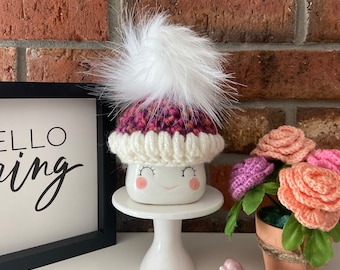 Könnte beinhalten: Eine weiße Keramiktasse mit einer rosa und lila gestrickten Mütze mit einem weißen Bommel. Die Tasse hat ein fröhliches Gesicht mit rosa Wangen. Die Tasse steht auf einem weißen Tortenständer. Im Hintergrund sind ein kleiner rosa Vogel und eine Topfpflanze mit rosa gehäkelten Blumen zu sehen.