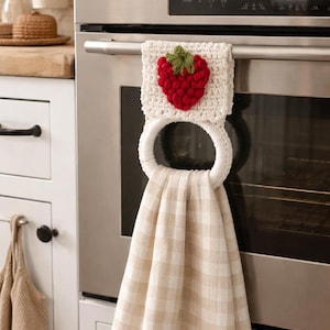 Puede incluir: Un pa&ntilde;o de cocina colgante con un patr&oacute;n de cuadros beige y blanco, sujeto a un soporte de anillo de ganchillo blanco. La parte superior del soporte est&aacute; decorada con una fresa de ganchillo roja tridimensional con hojas verdes.